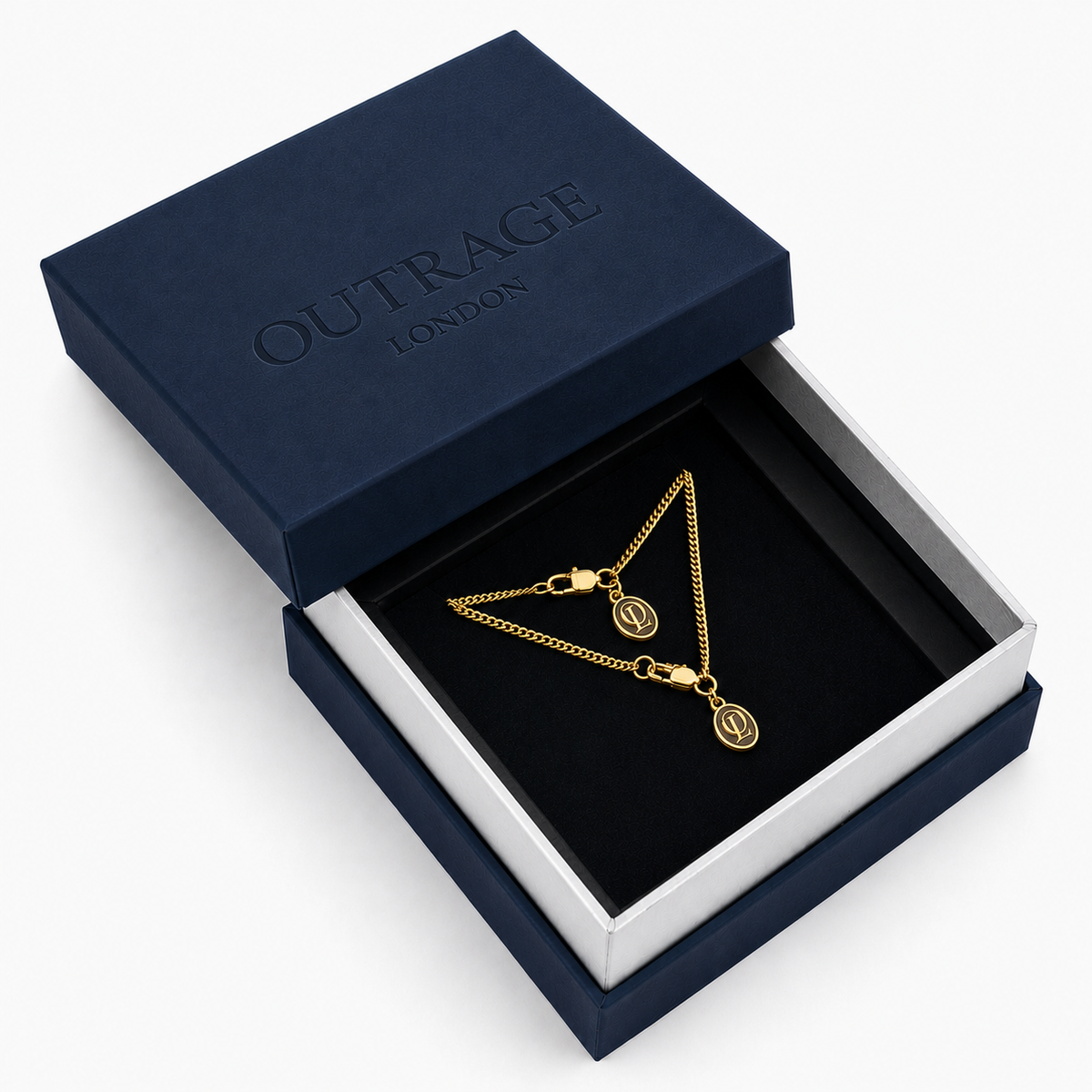 A gold chain necklace with two oval monogram pendants displayed inside an open navy blue Outrage London gift box on a white background.