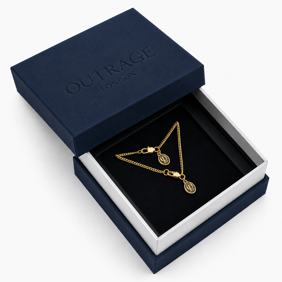 A gold chain necklace with two oval monogram pendants displayed inside an open navy blue Outrage London gift box on a white background.