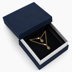A gold chain necklace with two oval monogram pendants displayed inside an open navy blue Outrage London gift box on a white background.