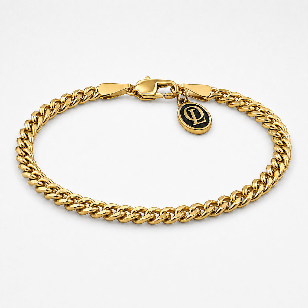 A gold-toned Cuban link chain bracelet with a lobster clasp and a small oval charm featuring a black monogram logo, shown on a light gray background.