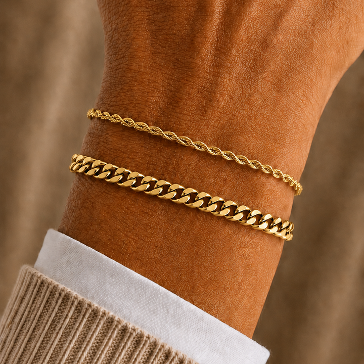 Two gold bracelets, one a delicate chain and the other a wider curb link style, worn on a person's wrist over a white shirt cuff and beige sweater.