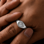 A silver signet ring with an engraved circular emblem on the finger, shown in a close-up lifestyle shot against warm skin tones.