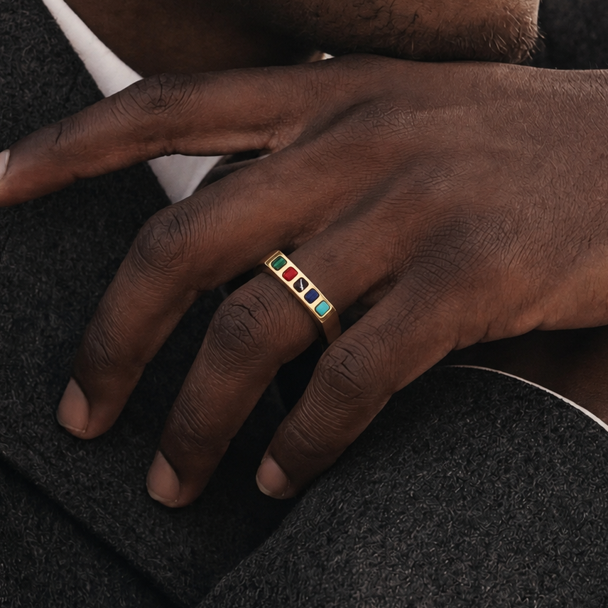 A close-up of a person's hand wearing a gold ring with four colorful rectangular inlays in red, green, blue, and teal, resting on dark textured fabric.