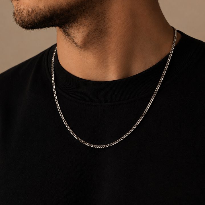 A close-up of a man's neck and lower face wearing a delicate silver chain necklace over a black crewneck shirt, set against a warm beige background.