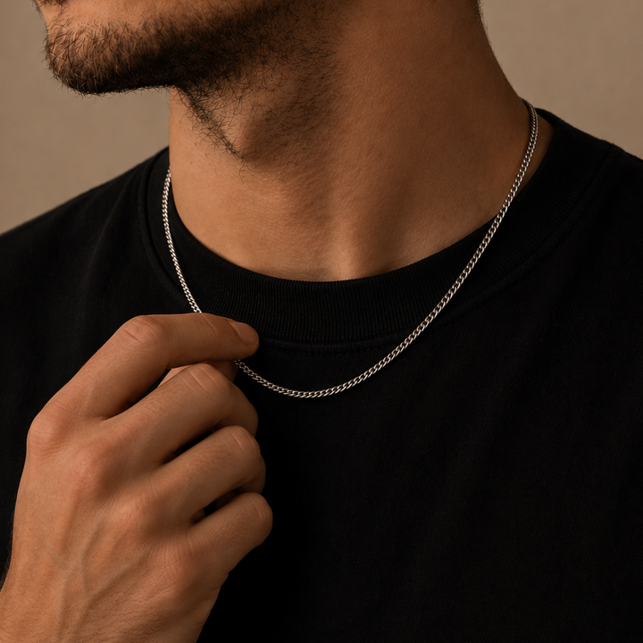 A close-up of a man wearing a delicate silver chain necklace over a black crewneck shirt, with his hand gently touching the chain near his collarbone.