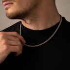 A close-up of a man wearing a silver chain necklace over a black crewneck sweatshirt, with his hand adjusting the chain.