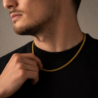 A close-up of a man wearing a gold chain necklace over a black crewneck shirt, with his hand resting near the chain.