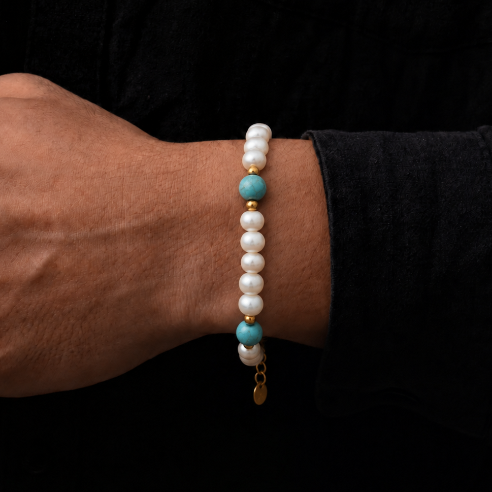 A close-up of a person's wrist wearing a beaded bracelet with white pearl-like beads, turquoise blue accent beads, and small gold spacer beads, set against a dark black shirt background.