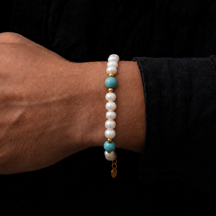 A close-up of a person's wrist wearing a beaded bracelet with white pearl-like beads, turquoise blue accent beads, and small gold spacer beads, set against a dark black shirt background.
