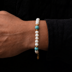 A close-up of a person's wrist wearing a beaded bracelet with white pearl-like beads, turquoise blue accent beads, and small gold spacer beads, set against a dark black shirt background.