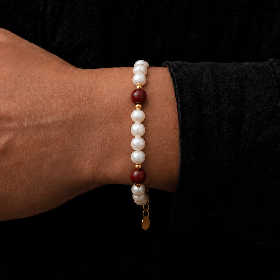 A beaded bracelet worn on a person's wrist, featuring white pearl-like beads, dark red stone beads, and small gold spacer rings with a dangling gold coin charm, set against a black background.