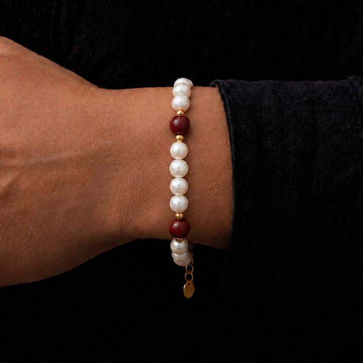 A beaded bracelet worn on a person's wrist, featuring white pearl-like beads, dark red stone beads, and small gold spacer rings with a dangling gold coin charm, set against a black background.
