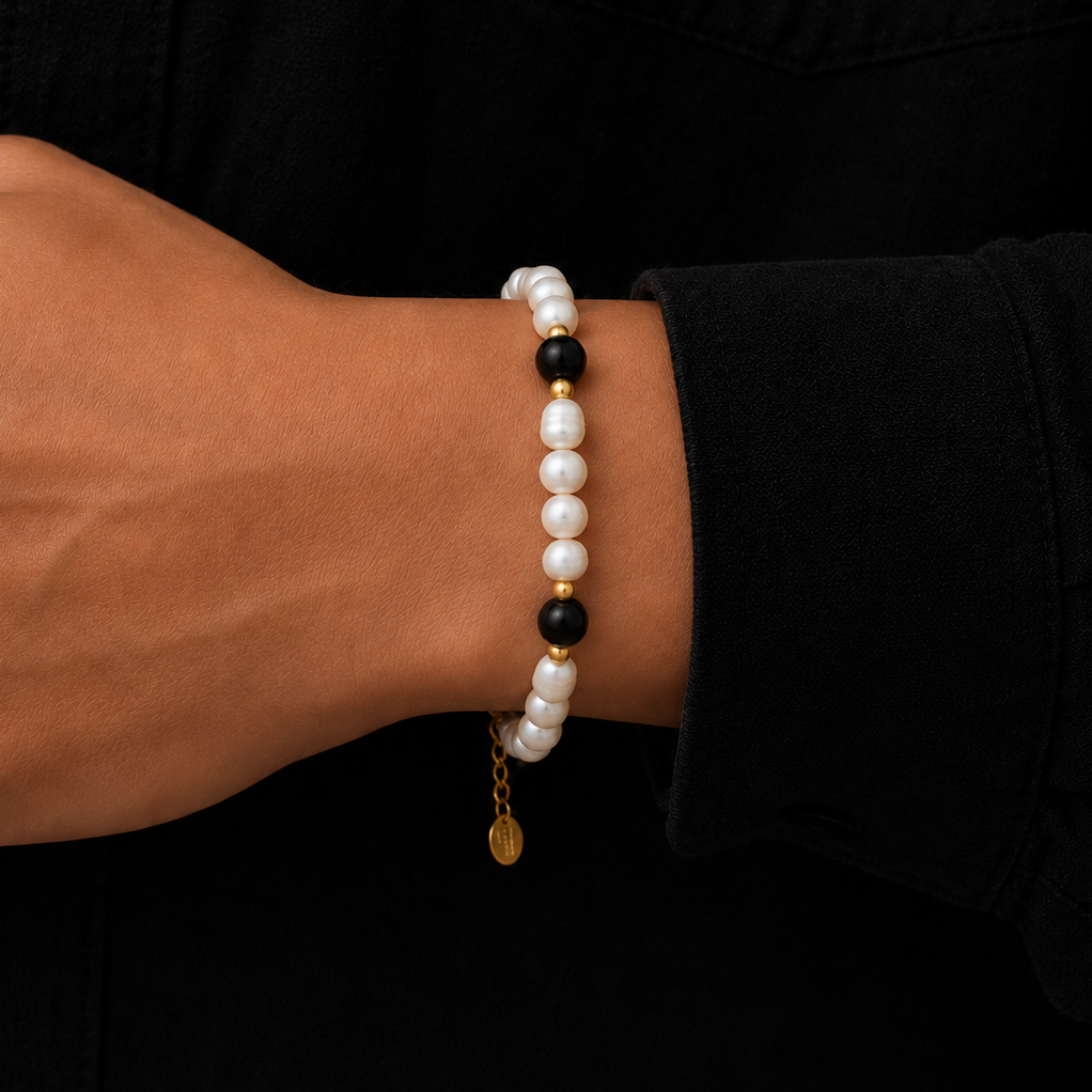 A close-up of a person's wrist wearing a beaded bracelet with white pearl-like beads, black beads, and small gold spacer beads, against a black shirt background.