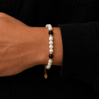 A close-up of a person's wrist wearing a beaded bracelet with white pearl-like beads, black beads, and small gold spacer beads, against a black shirt background.