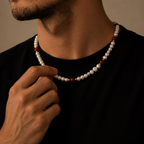 A close-up of a man wearing a beaded necklace with white pearls and small dark red beads, adjusting the necklace with his hand against a warm brown background.
