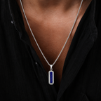 A close-up of a person wearing a silver chain necklace with a vertical rectangular pendant featuring a deep blue lapis lazuli stone in a silver bezel setting, worn over a black shirt.