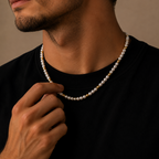 A close-up of a man wearing a delicate necklace made of white pearls and small gold beads, adjusting the chain with his hand against a warm beige background.