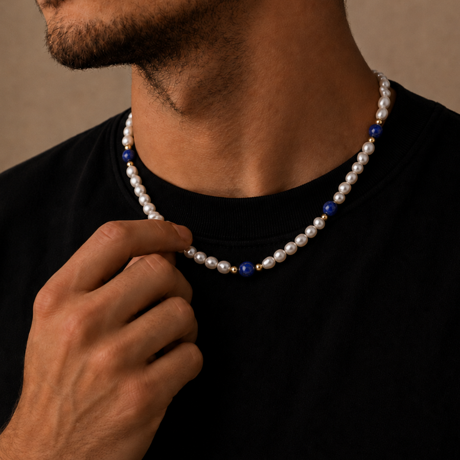 A close-up lifestyle shot of a man wearing a classic white pearl necklace with a black t-shirt, adjusting the necklace with his hand against a warm, neutral background.