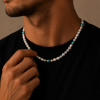 A close-up lifestyle shot of a man wearing a double-strand pearl necklace with small turquoise blue beads, adjusting the necklace with his hand against a warm brown background.