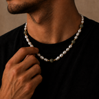 A close-up lifestyle shot of a man wearing a beaded necklace with white pearls, olive green beads, and small gold spacer rings, adjusting the necklace with his hand against a warm, textured beige background.
