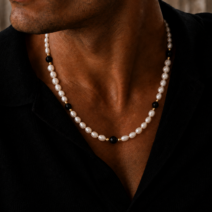 A close-up of a man wearing a pearl necklace with small black beads and gold spacer accents, styled with a black shirt.