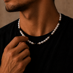 A close-up lifestyle shot of a man wearing a delicate white pearl necklace with small gold and dark metallic spacer beads, paired with a black t-shirt against a warm, textured brown background.