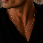 A close-up lifestyle shot of a man wearing a delicate silver chain necklace over a black shirt, with a blurred rustic wooden background.