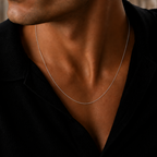 A close-up of a man wearing a delicate silver chain necklace over a black shirt, with a softly blurred stone background.