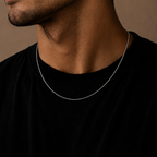 A close-up of a man's neck and lower face wearing a delicate silver chain necklace over a black t-shirt, set against a warm brown background.
