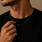 A close-up lifestyle shot of a man wearing a delicate silver chain necklace, touching the chain with his hand. He is wearing a black crew-neck t-shirt against a warm, textured beige background.