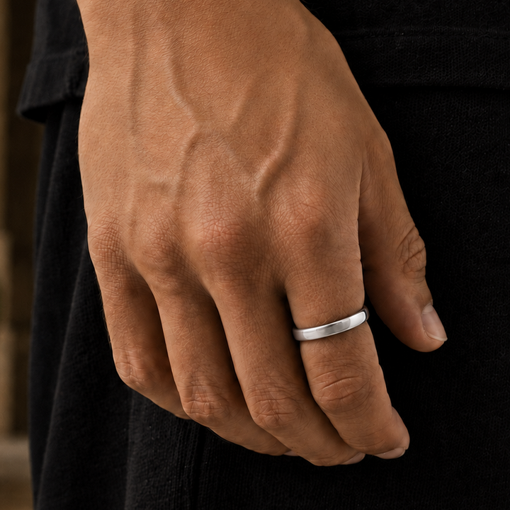 A close-up of a person's hand wearing a simple, polished silver band ring, set against a dark black outfit with a softly blurred outdoor background.