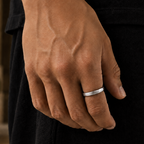 A close-up of a person's hand wearing a simple, polished silver band ring, set against a dark black outfit with a softly blurred outdoor background.