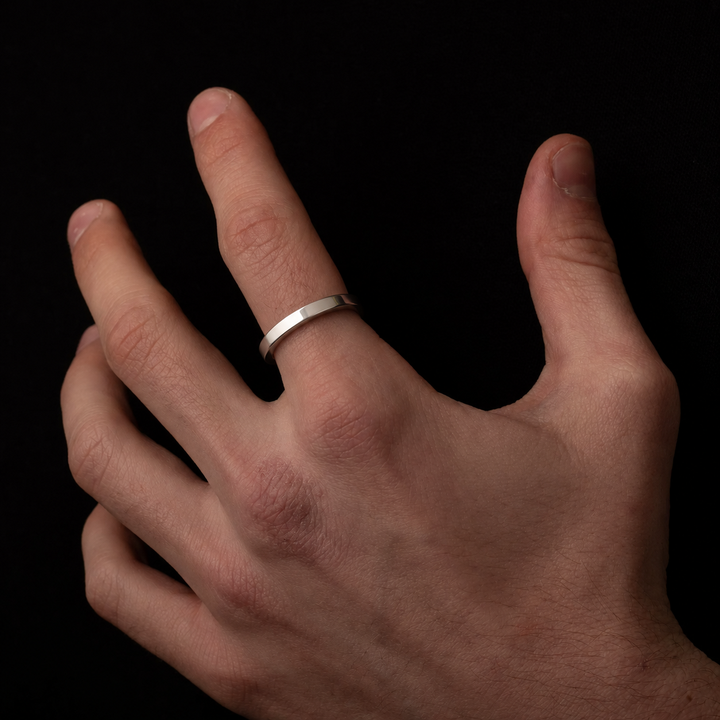 A minimalist silver band ring worn on the ring finger of a hand, shown against a solid black background.