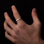 A minimalist silver band ring worn on the ring finger of a hand, shown against a solid black background.