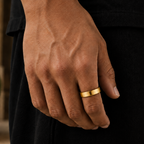 A close-up of a person's hand wearing a simple, wide gold band ring, dressed in a black shirt and black trousers, with a softly blurred outdoor background.