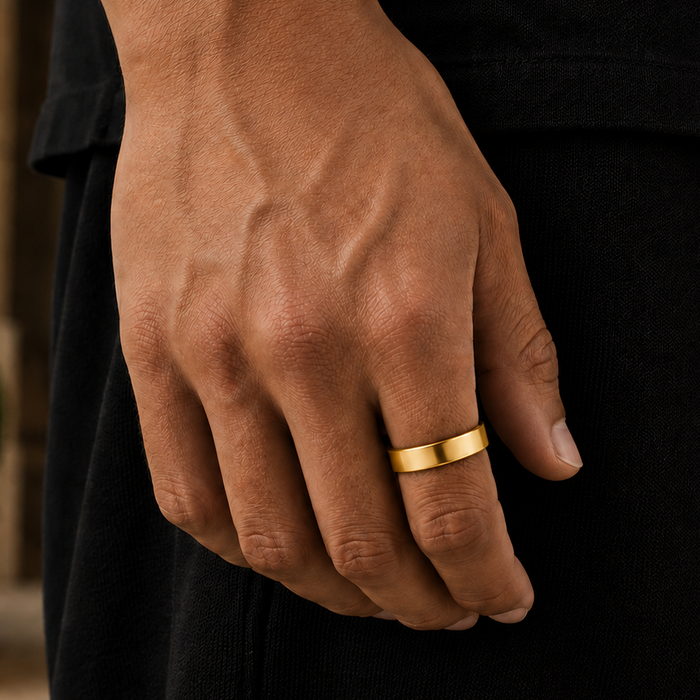 A close-up of a person's hand wearing a simple, wide gold band ring, dressed in a black shirt and black trousers, with a softly blurred outdoor background.