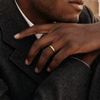 A close-up of a man's hand resting on his lap, wearing a simple, polished gold band ring. He is dressed in a dark charcoal gray wool blazer over a white collared shirt.
