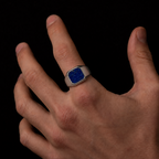 A silver signet ring with a square blue lapis lazuli stone inlay, shown worn on a person's ring finger against a solid black background.