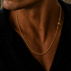 A close-up of a man wearing a delicate gold chain necklace with a small charm, styled with a black shirt against a rustic wooden background.