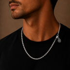 A close-up of a man's neck and lower face wearing a silver chain necklace with a small circular charm, over a black t-shirt against a warm brown background.