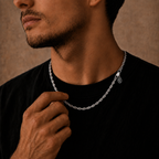 A close-up lifestyle shot of a man wearing a silver chain necklace with a small hanging monogram pendant, styled with a black t-shirt against a textured brown background.