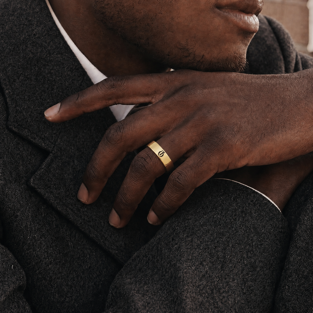 A close-up lifestyle shot of a man wearing a slim gold band ring with a small engraved symbol, resting his hand on his lap over a dark gray wool coat.