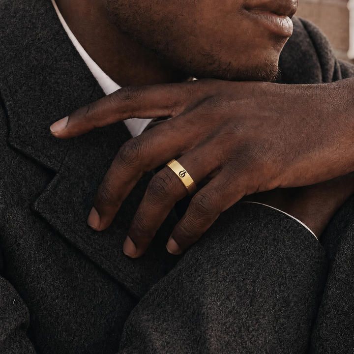 A close-up lifestyle shot of a man wearing a slim gold band ring with a small engraved symbol, resting his hand on his lap over a dark gray wool coat.