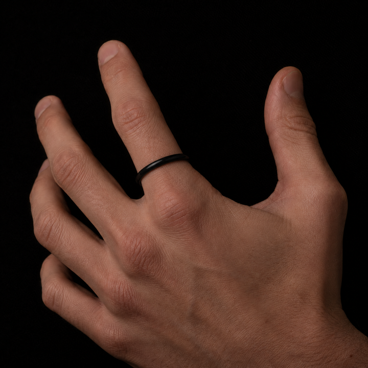 A minimalist black metal band ring worn on the ring finger of a hand, shown against a solid black background.