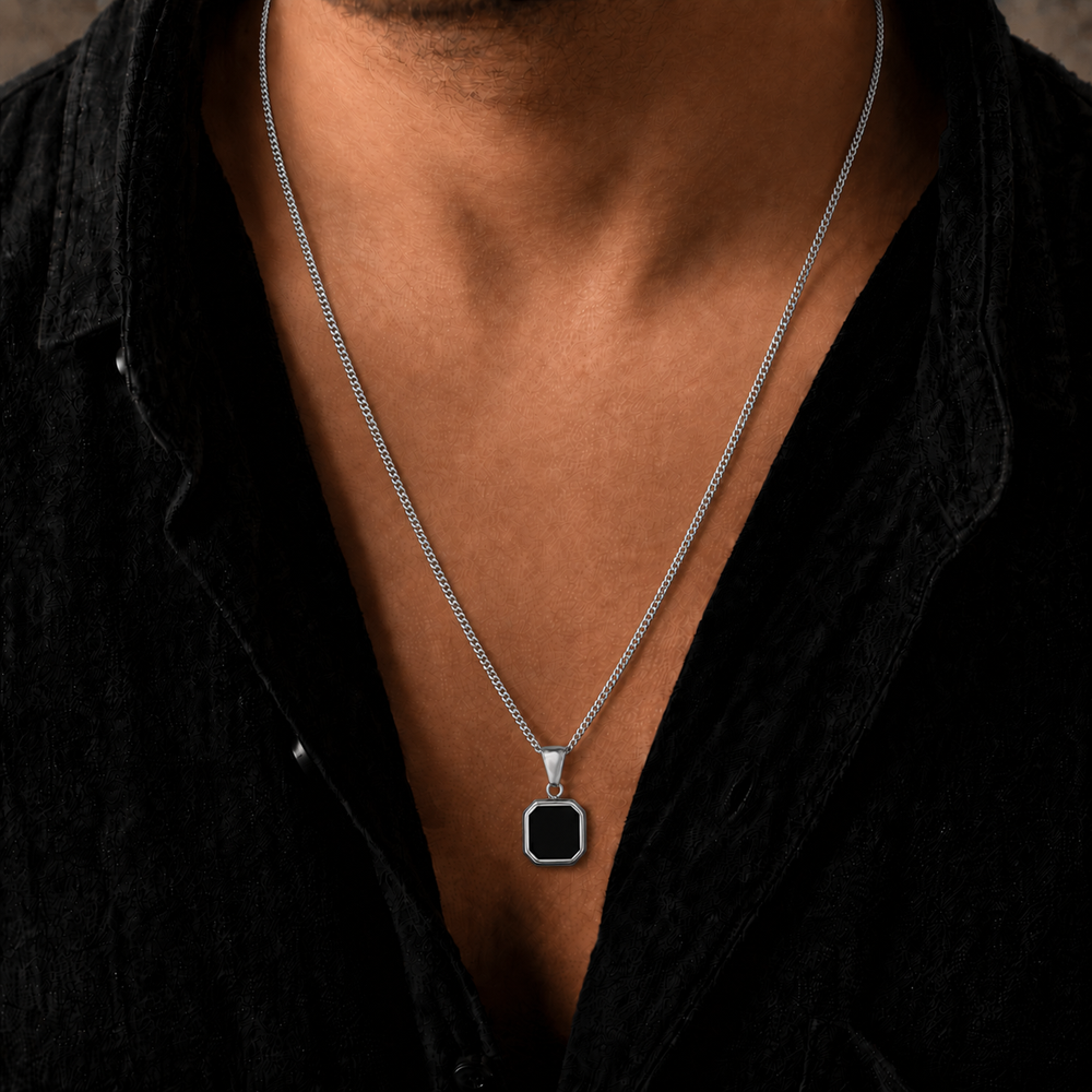 A silver-toned chain necklace with a square black pendant, worn by a man in a black shirt.