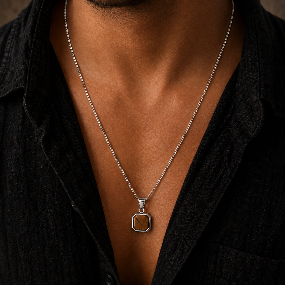 A close-up of a man wearing a silver chain necklace with a square pendant featuring a brown textured stone inlay, styled with a black shirt.