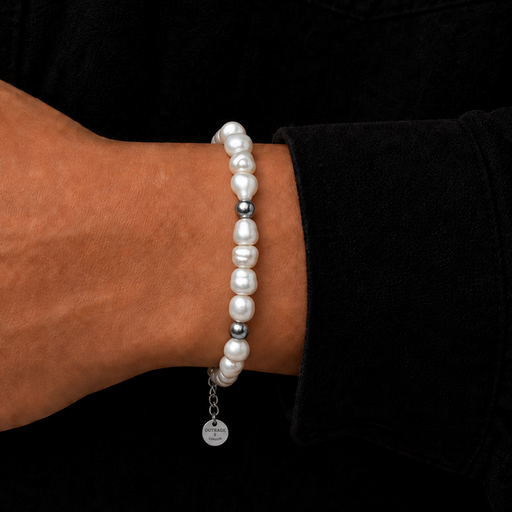 A close-up of a person's wrist wearing a beaded bracelet made of white pearls and small silver spacer beads, with a small silver charm hanging from the end, set against a black shirt background.