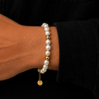 A close-up of a person's wrist wearing two stacked bracelets: one strand of white freshwater pearls with gold spacer beads, and another pearl bracelet with a small gold disc charm engraved with 'LOVE YOURSELF'.