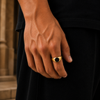 A close-up of a person's hand wearing a gold signet ring with a black oval center, set against a dark shirt and blurred stone background.