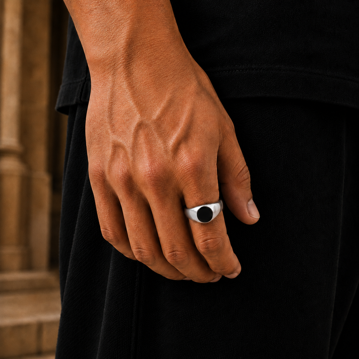 A close-up of a man's hand wearing a silver signet ring with a black oval face, set against a dark shirt and blurred stone background.
